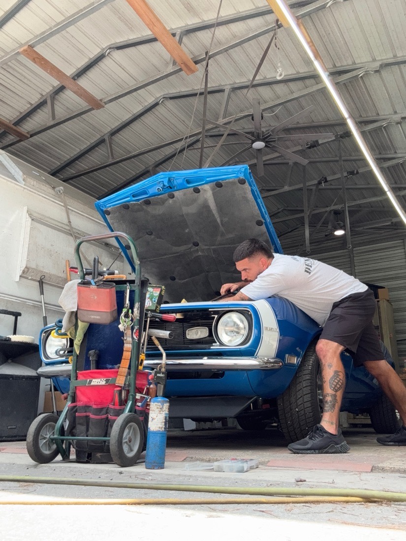 A Gold Coast PDR technician reaching over the front fender of a blue and white first-generation Camaro with the hood raised.