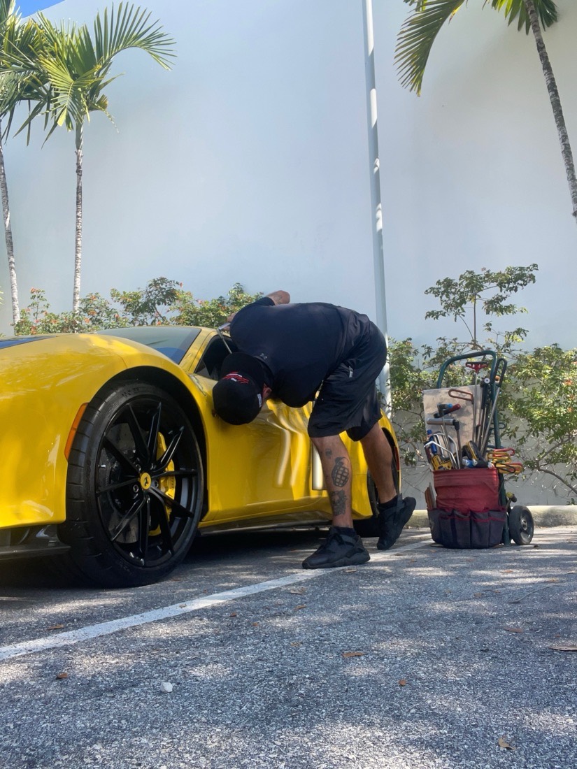 A Gold Coast PDR technician leaning over the door of a yellow Ferrari in a parking lot, tool cart parked on the asphalt beside it.