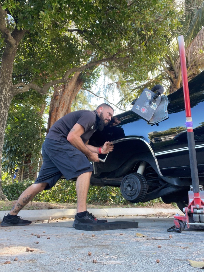 A Gold Coast PDR technician working a rear quarter panel on a black classic raised on a jack, outdoors under the trees.