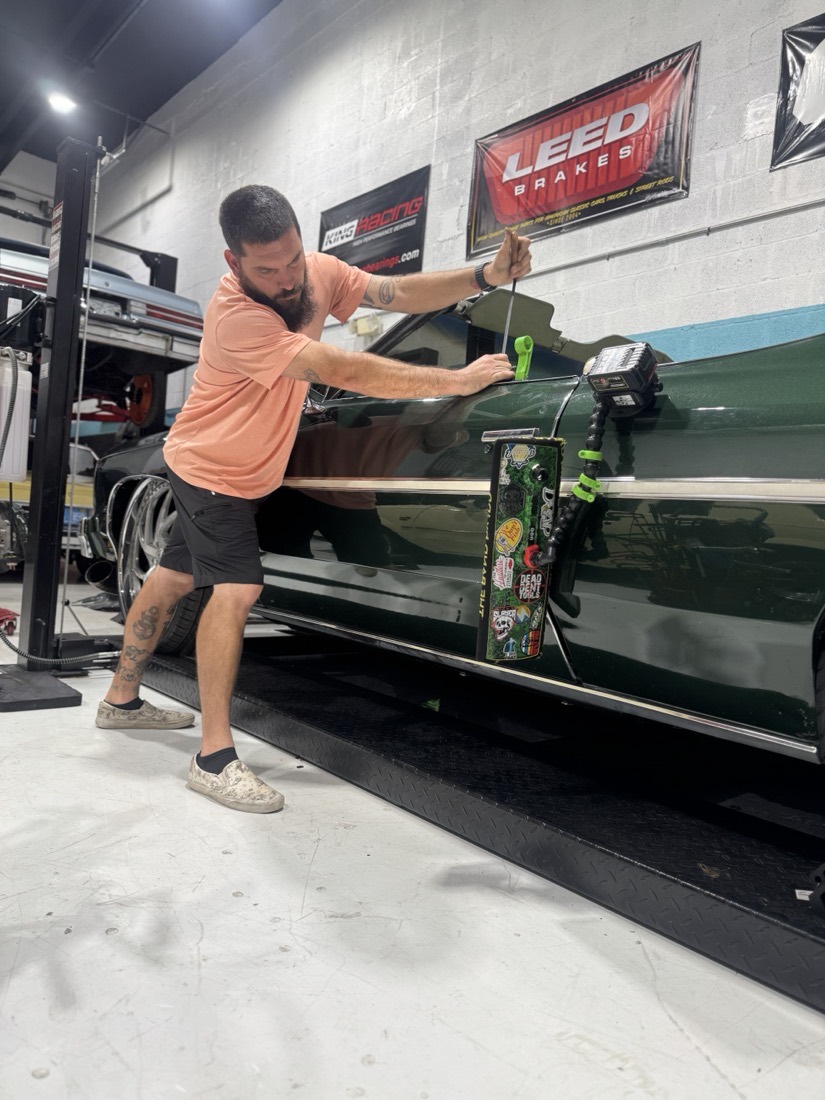A Gold Coast PDR technician braced against a dark green classic, working a rod along the body line under the shop lights.
